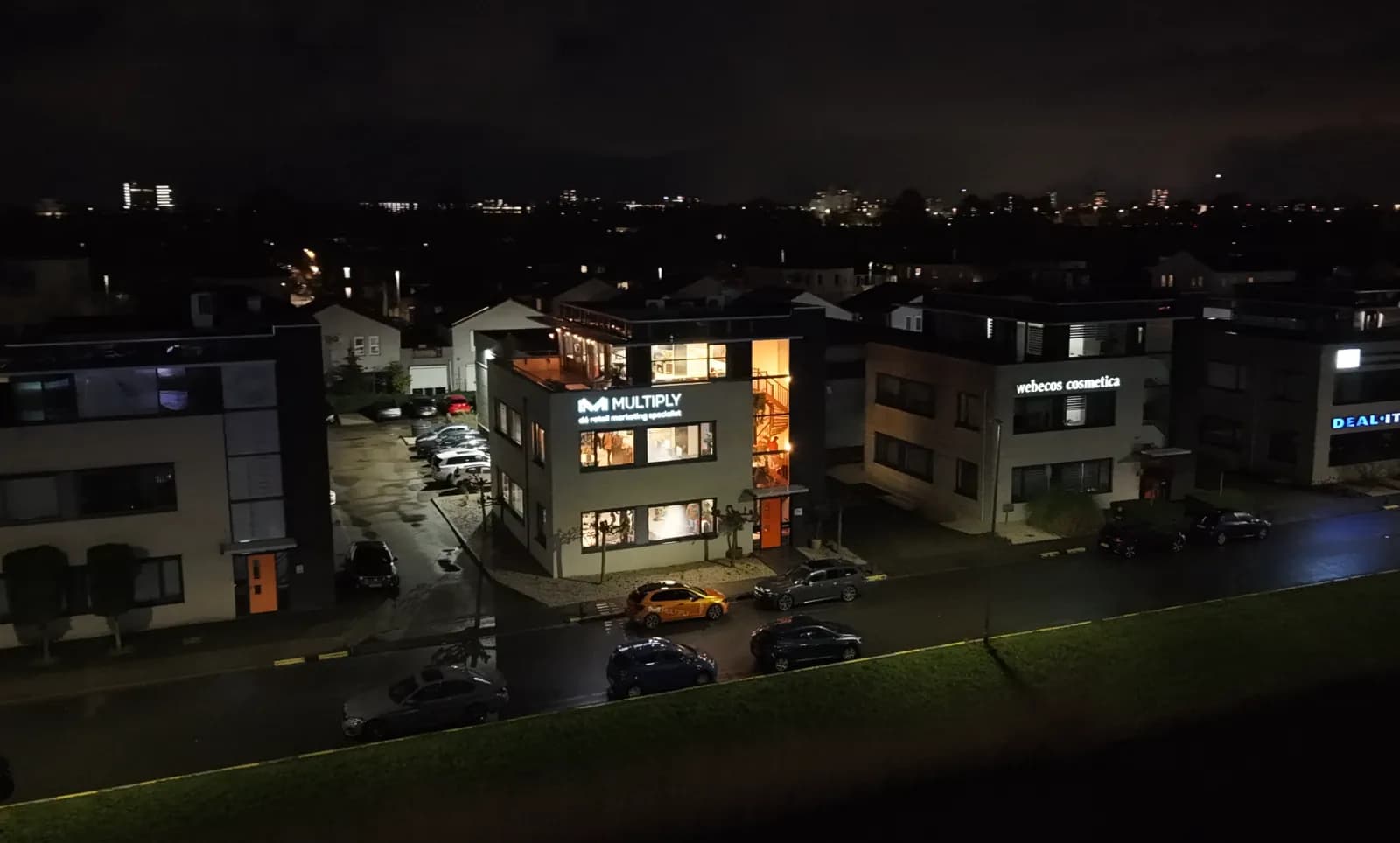 Het Multiply-kantoor in de avond, met verlicht logo en de oranje Multiply-auto voor de deur